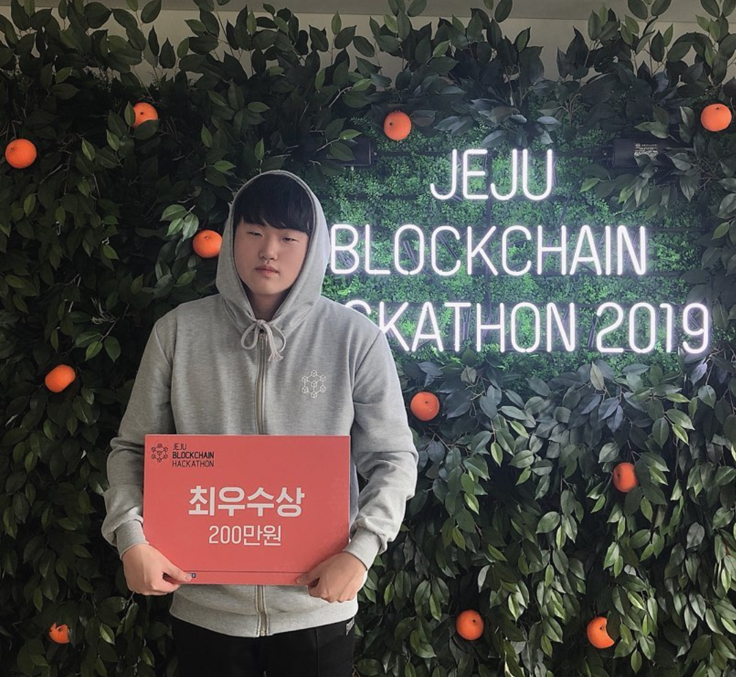 JEJU BLOCKCHAIN HACKATHON 2019 네온사인과 귤이 달린 초록 벽 앞에서, 회색 후드를 입고 '최우수상 200만원'이라고 적힌 빨간 상패를 들고 서 있는 나.